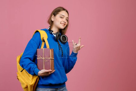 Front view of pretty brunette schoolgirl standing, showing rock. Young trendy student wearing fashionable clothes, looking at camera, smiling. Concept of youth and education.