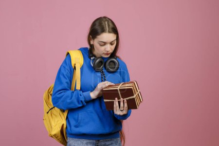 Front view of pretty brunette girl standing, holding bunch of old books. Beautiful schoolgirl studying, looking down, holding yellow rucksack. Isolated on pink studio background.