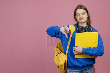Front view of young female student standing, showing dislike. Pretty schoolgirl looking at camera, gloomy, holding yellow rucksack and folder, hating school. Concept of youth.