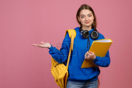 Front view of pretty brunette schoolgirl standing, holding yellow backpack and folder. Beautiful young female looking at camera, smiling. Isolated on pink studio background.