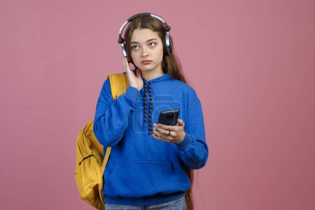 Front view of young pretty female standing, holding, using smartphone. Beautiful schoolgirl with earphones, looking forward, holding yellow rucksack. Isolated on pink background.