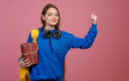 Front view of brunette schoolgirl standing, raising hand, strong. Pretty young student with long hair and earphones looking at camera, smiling. Isolated on pink studio background.