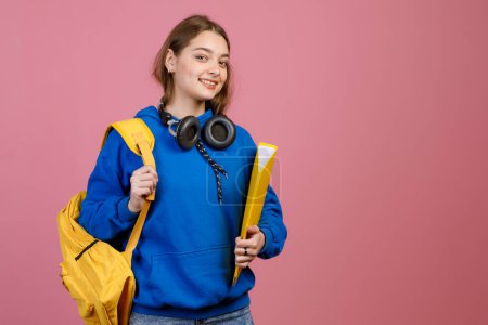 Front view of young female student holding yellow rucksack and folder, smiling. Pretty schooolgirl with long hair, looking at camera indoors. Isolated on pink studio background.
