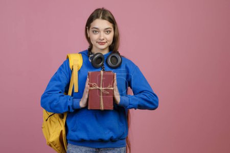 Front view of pretty schoolgirl holding bunch ob old books, showing by hands. Young brunette woman with earphones, standing, looking at camera, smiling. Concept of youth.