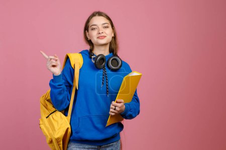 Front view of pretty brunette female standing, pointing by finger. Beautiful schoolgirl looking at camera, smiling, holding yellow rucksack and folder. Concept of youth life.