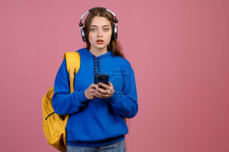 Front view of brunette schoolgirl standing, looking ar camera with open mouth. Pretty young female using smartphone, listening to music in earphones, surprised. Concept of youth.