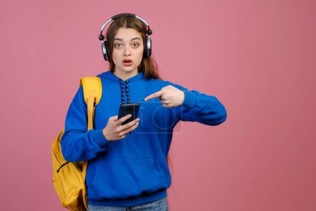 Front view of slim schoolgirl standing, using smartphone, pointing by finger. Beautiful young female looking at camera with open mouth, surprised. Isolated on pink background.