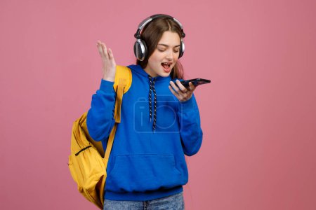 Front view of pretty brunette schoolgirl standing, using smartphone, calling, taking, crying. Beautiful young student looking down, smiling. Isolated on pink studio background.