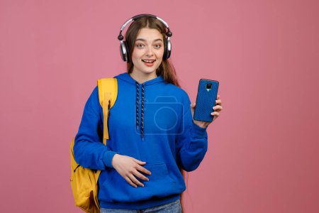 Front view of surprised, glad, happy girl standing, showing smartphone. Pretty slim student with long hair looking at camera, listening to msuic in earphones. Concept of youth.