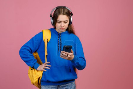 Front view of frown schoolgirl standing, using smartphone, looking. Young pretty woman with long hair holding hand on waist, using earphones. Isolated on pink studio background.