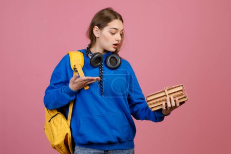Front view of trendy schoolgirl standing, holding bunch of old books. Young brunette female looking at books, surprised, asking, with earphones on neck. Concept of youth life.