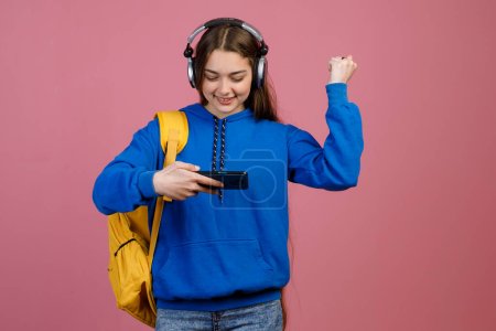 Front view of brunette schoolgirl using smartphone, watching. Pretty young female with long hair raising hand, showing hurray, glad, happy. Isolated on pink studio background.