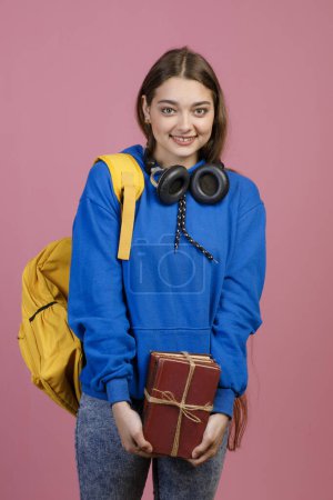 Front view of brunette schoolgirl standing, looking at camera, smiling. Trendy young student smiling, holding bunch of books and rucksack. Isolated on pink studio background.