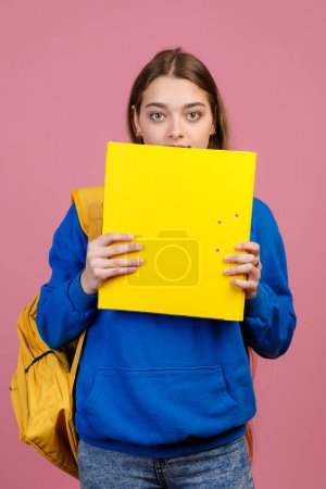 Front view of surprised young female standing, closing mouth with big yellow folder, holding by hands. Pretty schoolgirl looking at camera. Isolated on pink studio background.