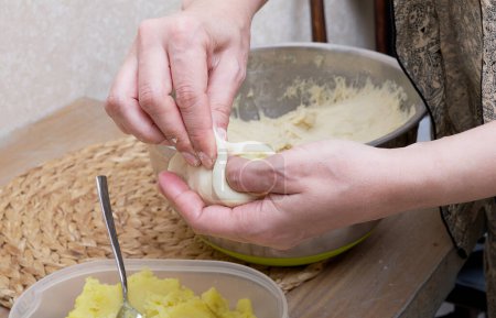 Téléchargez les photos : Les mains des femmes tiennent soigneusement une purée de pomme de terre l'enveloppant dans la pâte chaude dans la cuisine de la maison sur la table en bois de la ferme confortable. Le processus de préparation à la main d'un gros plan de tarte. - en image libre de droit