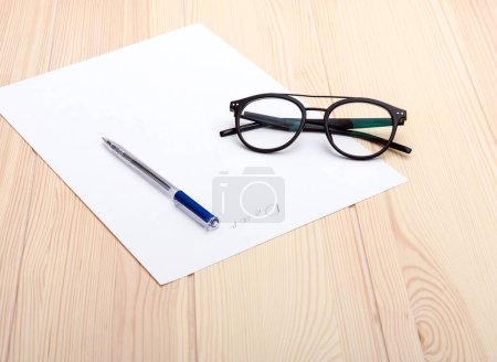 Photo for Exquisite black glasses are arranged on a light wood structural craft and texture office table next to a sheet of white paper with a blue pen on the smooth surface of the paper. Studio photo. - Royalty Free Image