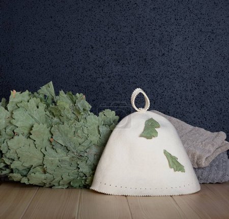 Téléchargez les photos : Photos of a sauna in the Finnish style where a special hat for a bath, a broom made of oak leaves and a stack of large gray towels lie on a wooden textured table. - en image libre de droit