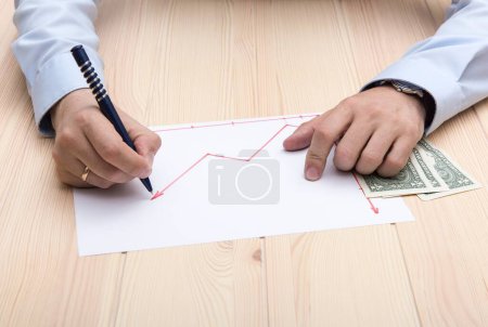 Photo for The hands of a man dressed on blue office t-shirt with classic wrist watch and golden ring on his hands, draws using red pencil a chart of rising stock prices on the rustic wooden structure table. - Royalty Free Image
