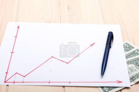 Photo for A drawing of a stock price growth chart on a table made with a red pen placed on the surface of a light wooden office table. A banknote lies under a sheet of paper. - Royalty Free Image