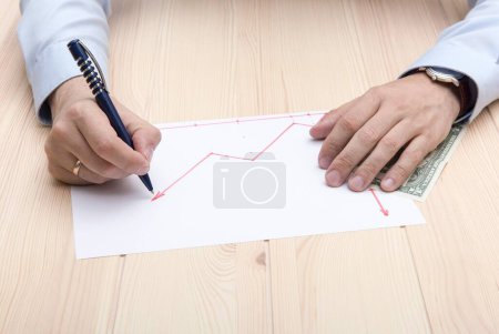 Photo for The hands of a man in a close-up, dressed in a blue office shirt with a classic wristwatch and a gold ring on his hands, draw a graph of the growth of stock prices on a sheet of white paper. - Royalty Free Image