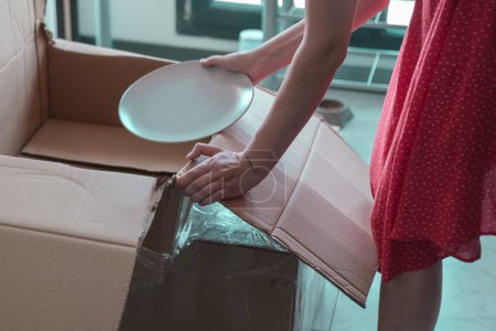 Photo for Young woman packing or unpacking kitchen stuff from the carton boxes after before moving, SSTK Home - Royalty Free Image