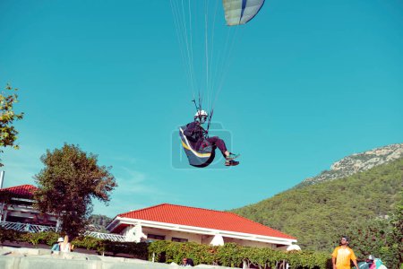 Foto de Mugla, fethiye, oludeniz - 23 de octubre de 2022: piloto de parapente aterrizando desde el cielo a una zona concurrida con casa, montañas, personas - Imagen libre de derechos