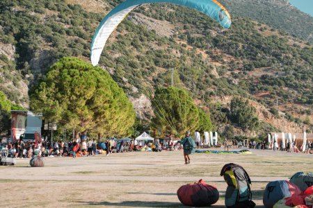 Foto de Mugla, fethiye, oludeniz - 23 de octubre de 2022: piloto de parapente aterrizando desde el cielo a una zona concurrida con casa, montañas, personas - Imagen libre de derechos