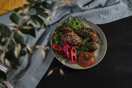 baked chicken wings and legs with baby rocca leaves, red pepper, homemade hot sauce dressed with sesame seeds and rosemarine, dark backround, blue textile cloth