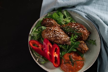 baked chicken wings and legs with baby rocca leaves, red pepper, homemade hot sauce dressed with sesame seeds and rosemarine, dark backround, blue textile cloth
