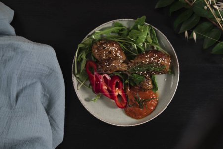 gebackene Hühnerflügel und -beine mit Baby-Rocca-Blättern, Paprika, hausgemachte scharfe Sauce mit Sesam und Rosmarin, dunklem Hintergrund, blauem Textiltuch