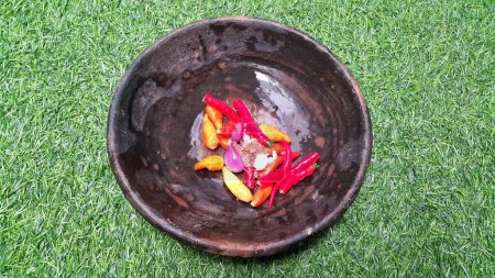 Foto de Close up of ingredients for making chili sauce on a clay plate. Red chili, onion, and salt. Onion and chili in a pestle and stone mortar. Cooking ingredients. Artificial green grass. - Imagen libre de derechos