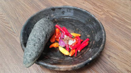 Foto de Close up of ingredients for making chili sauce on a clay plate. Red chili, onion, and salt. Onion and chili in a pestle and stone mortar. Cooking ingredients. Wooden background. - Imagen libre de derechos