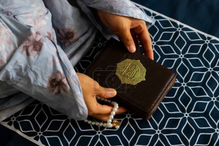 Jakarta, Indonesia - February 21, 2024: A Muslim woman sitting on a prayer mat and holding the quran with Indonesian translation