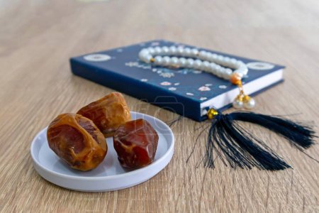 Blick auf einen Teller mit Dattelfrüchten auf dem Holztisch mit einem kleinen Buch und Gebetsperlen