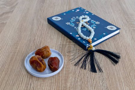 A plate of dates fruit on the wooden table with a small book and prayer beads