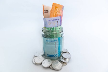 Billetes de Rupia indonesia en frasco de vidrio y monedas dispersas sobre fondo blanco