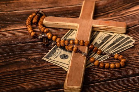 A Catholic cross, a rosary with beads and dollars lie on a dark brown wooden table.