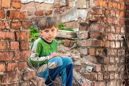 Photo for Poor and unhappy orphan boy, sitting on the ruins and ruins of a destroyed building. Staged photo. - Royalty Free Image