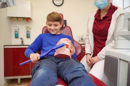 Téléchargez les photos : Un garçon dans le cabinet d'un dentiste tient une grande brosse à dents et une mâchoire en plastique dans ses mains. - en image libre de droit