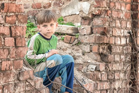 Poor and unhappy orphan boy, sitting on the ruins and ruins of a destroyed building. Staged photo.