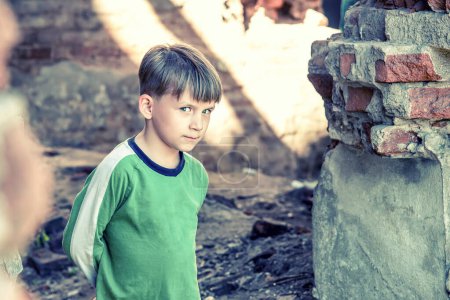 Poor and unhappy orphan boy, stands in a ruined building and looks out with danger. Staged photo.