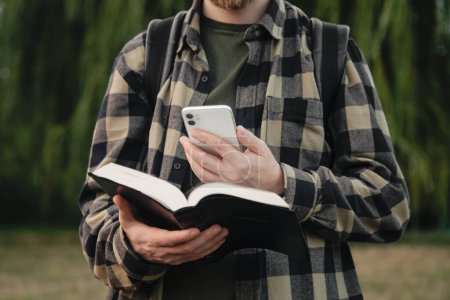 Photo for A man with a smartphone and a Bible in his hands outside, christian online technology concept, online live church, Holy Bible book and online study. - Royalty Free Image
