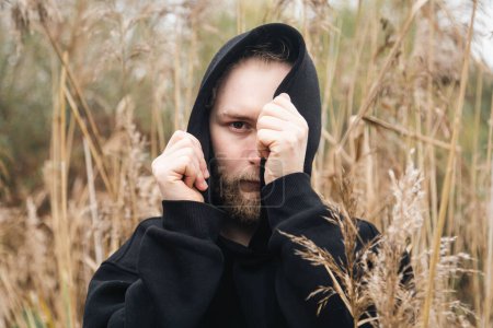 A young bearded man hides his face in a black hood, standing in the coastal reeds, the concept of mental health, loneliness and depression.
