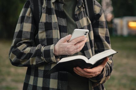 Photo for A man with a smartphone and a Bible in his hands outside, christian online technology concept, online live church, Holy Bible book and online study. - Royalty Free Image