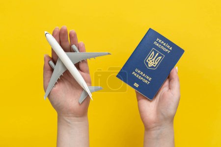 Photo for Ukrainian biometric passport and airplane model in female hands on a yellow background. The concept of tourism, travel and migration from Ukraine. - Royalty Free Image