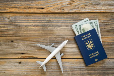 Photo for Ukrainian biometric passport, dollar bills and airplane figurine on a wooden background, top view. The concept of travel, migration and refugee from Ukraine. Copy space. - Royalty Free Image