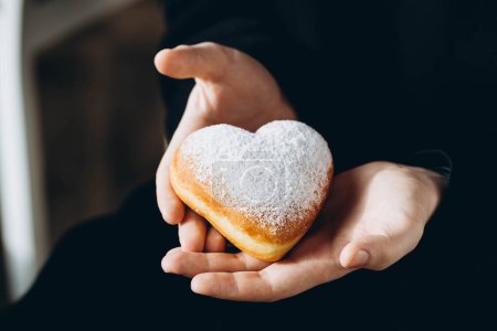 A man in a black hoodie holds a heart-shaped donut covered in powdered sugar, close up. Delicious snack, coffee break at work, lifestyle.