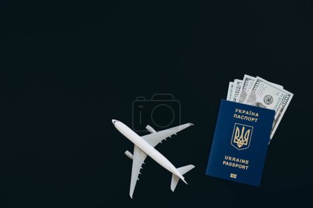 Ukrainian biometric passport, dollar bills and airplane figurine on a black background, flat lay. The concept of travel, migration and refugee from Ukraine. Copy space.