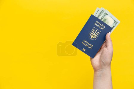 Biometric Ukrainian passport and money in a female hand on a yellow background isolated. Passport of a citizen of Ukraine and dollar bills. Copy space.
