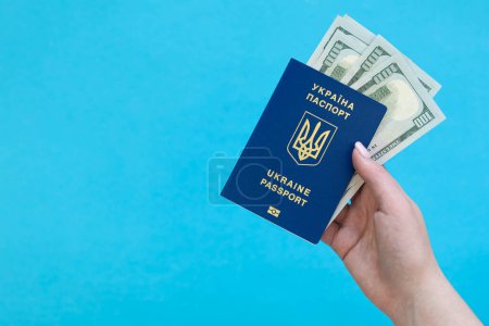 Biometric Ukrainian passport and money in a female hand on a blue background isolated. Passport of a citizen of Ukraine and dollar bills. Copy space.